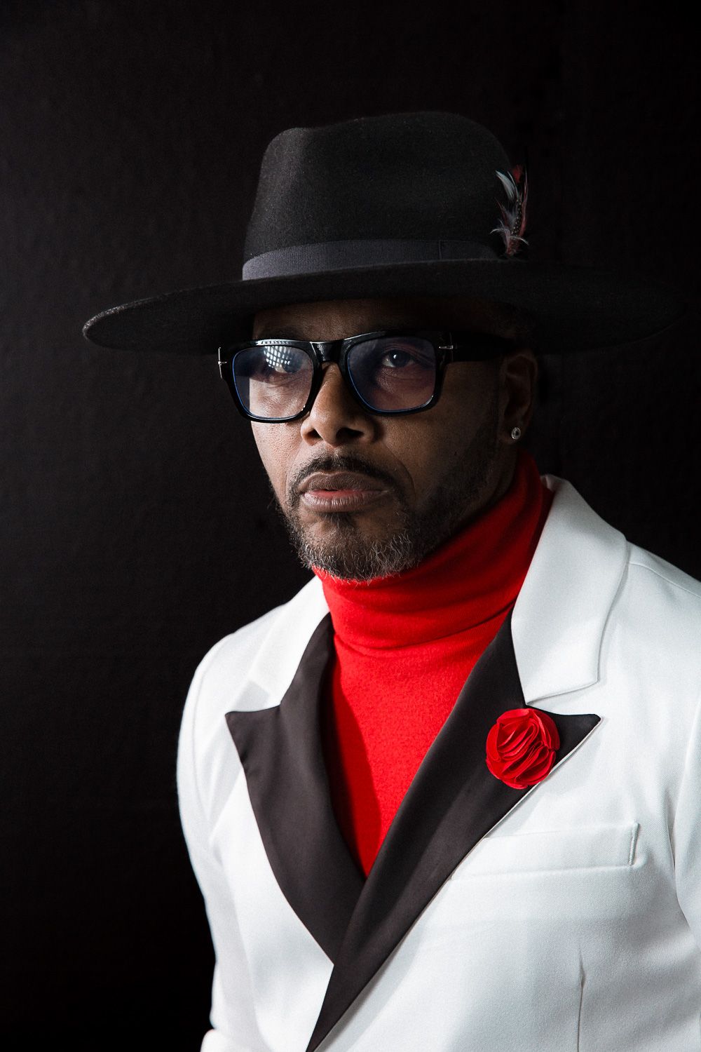 Portrait in white suit, red turtleneck, and black hat against dark background.