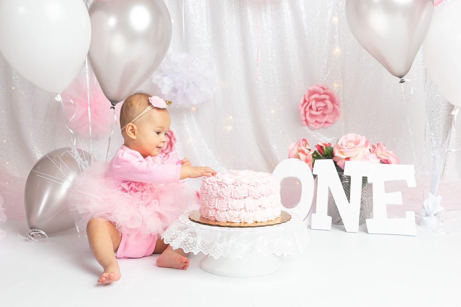 Baby girl in pink tutu sits beside a first birthday smash cake surrounded by balloons and ONE letters.