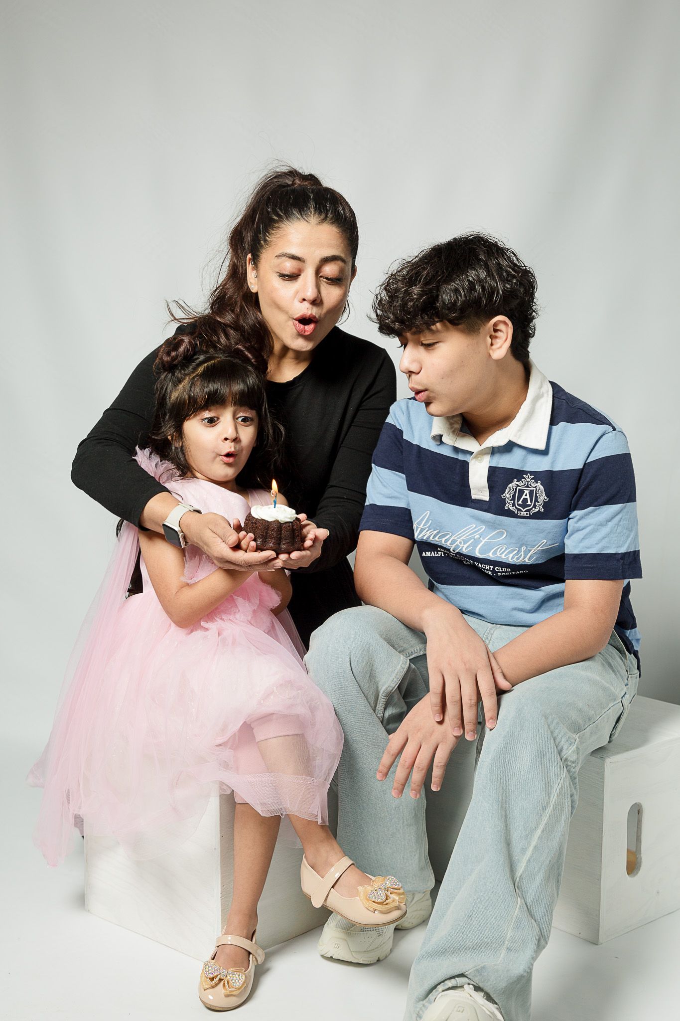 Family celebrating birthday with young girl in pink dress blowing out candle on cake with mother and brother.