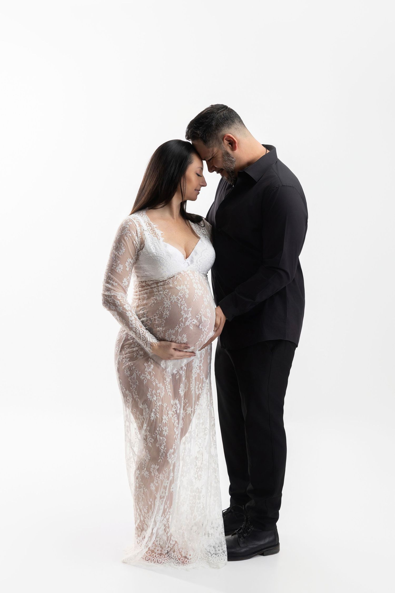 Romantic maternity photoshoot featuring couple in white lace dress and black outfit against white backdrop.