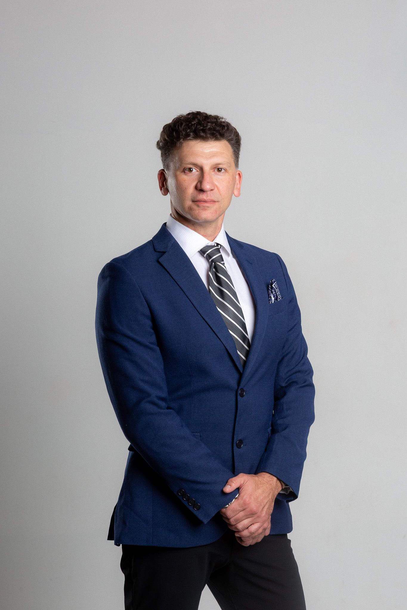 Professional wearing a navy blue suit and striped tie poses against light gray background.