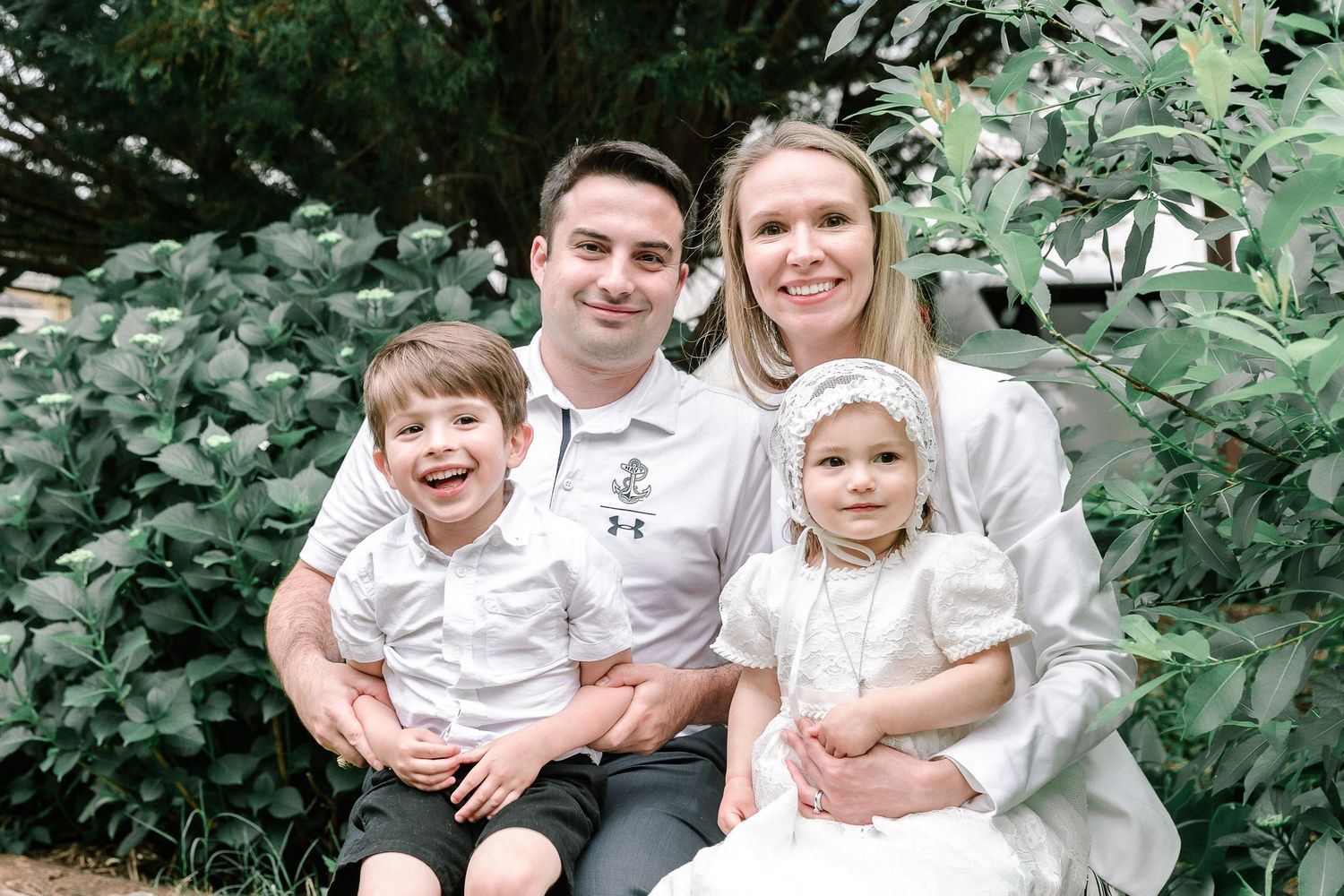 Family portrait in white and neutral clothing against lush green foliage background.
