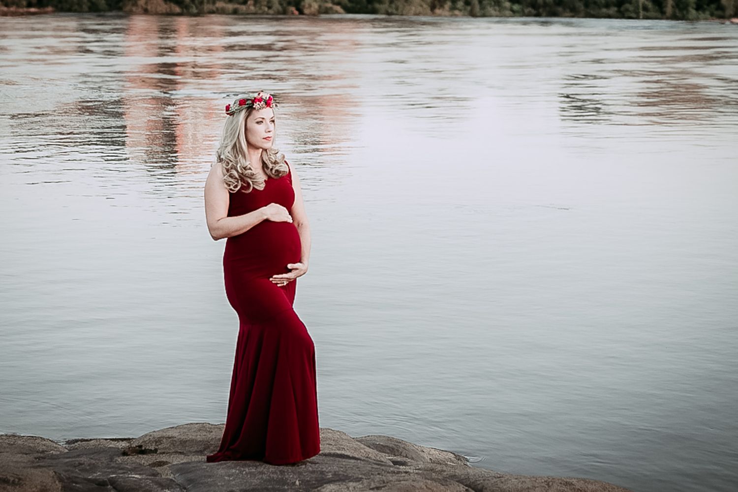 Maternity photo series in flowing red dress by tranquil waterfront.