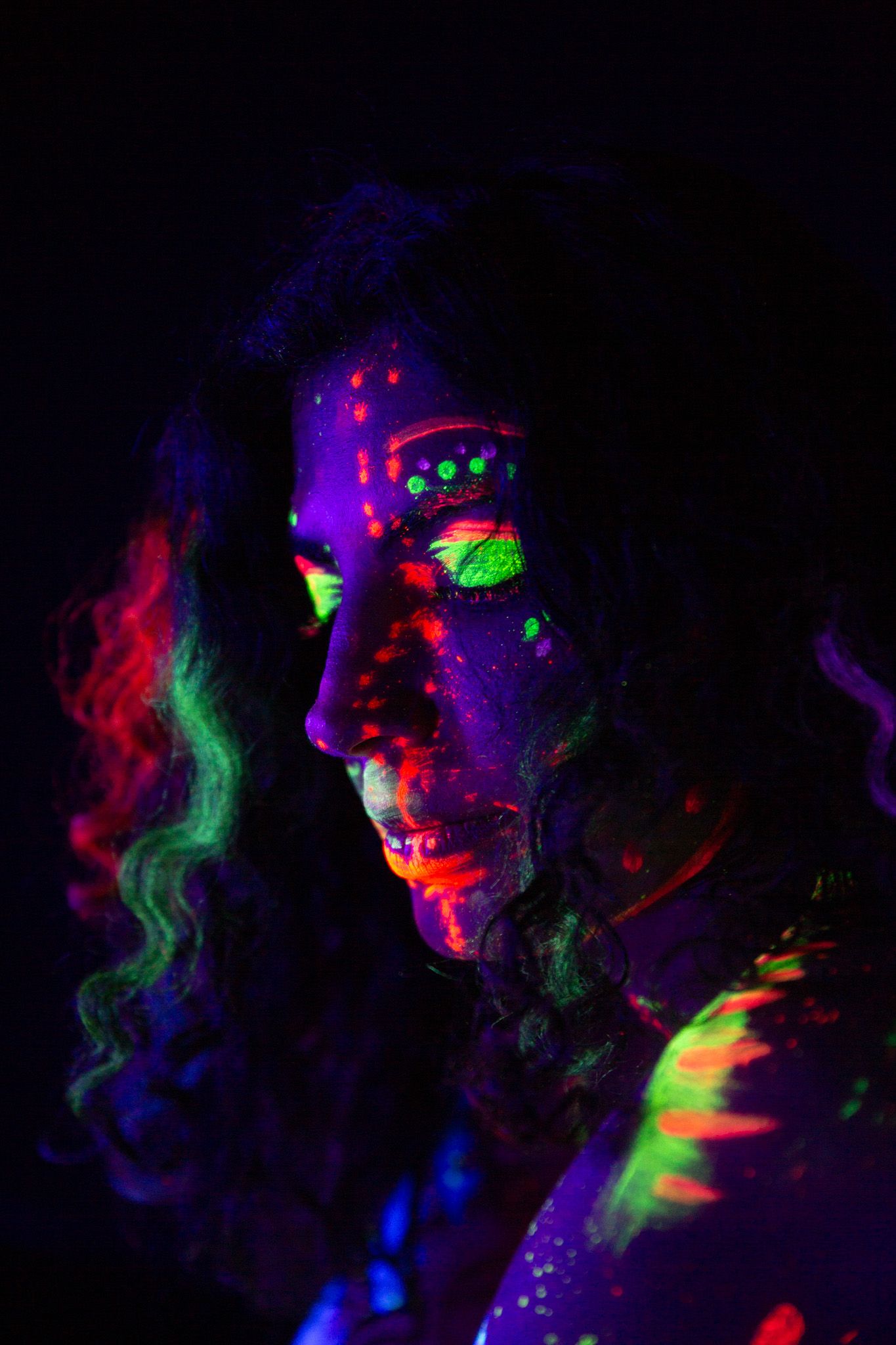 Close-up portrait with vibrant UV reactive face paint in neon colors against dark background.