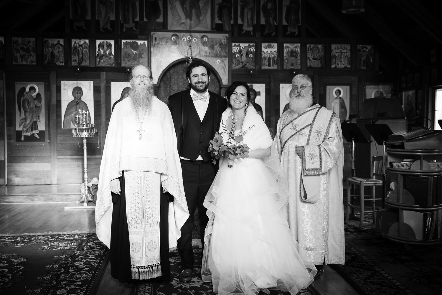 Black and white portrait of Orthodox wedding couple posing with two priests inside icon-adorned church.