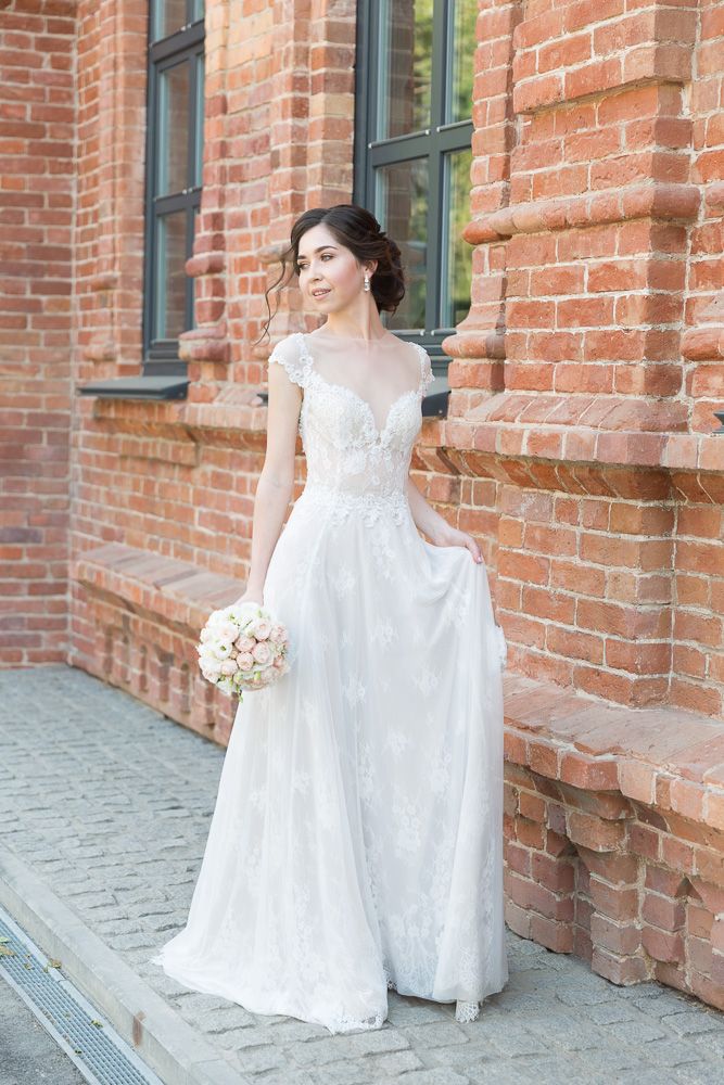 Elegant bride in lace wedding gown holding bouquet beside a red brick building outdoors.