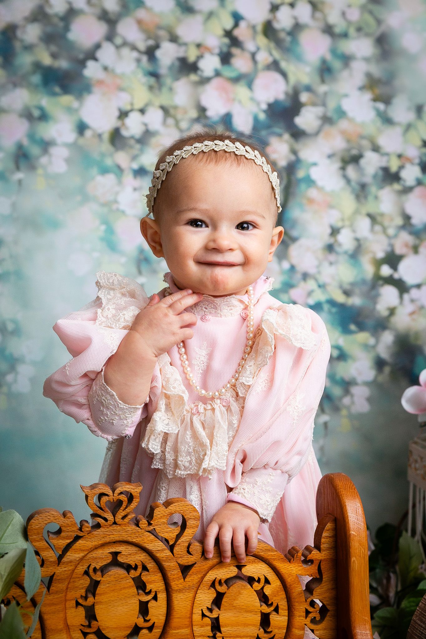 Smiling baby girl in pink lace dress with pearl necklace and headband, posing by ornate wooden furniture.