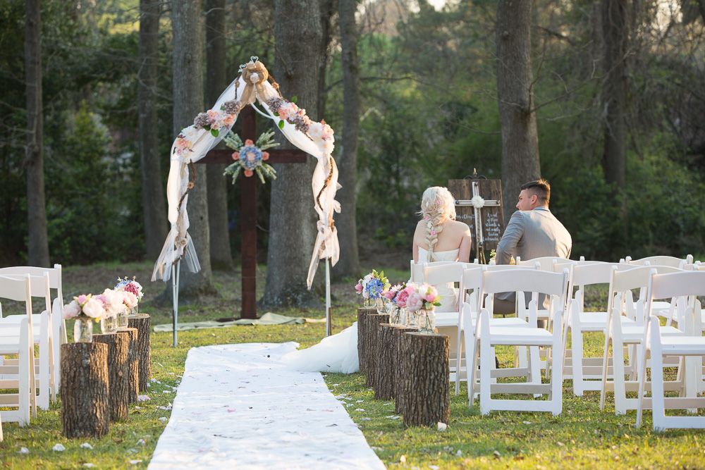 Elegant outdoor wedding ceremony with floral arch, white chairs, and tree stump aisle decor.