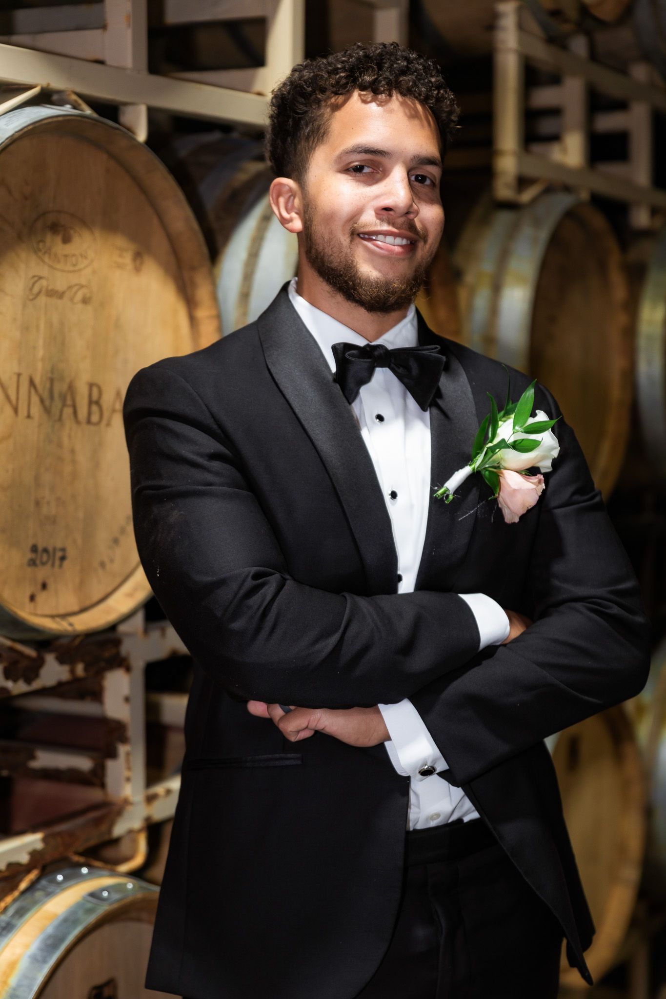 Young man in black tuxedo with boutonniere poses confidently in front of wine barrels in a cellar.