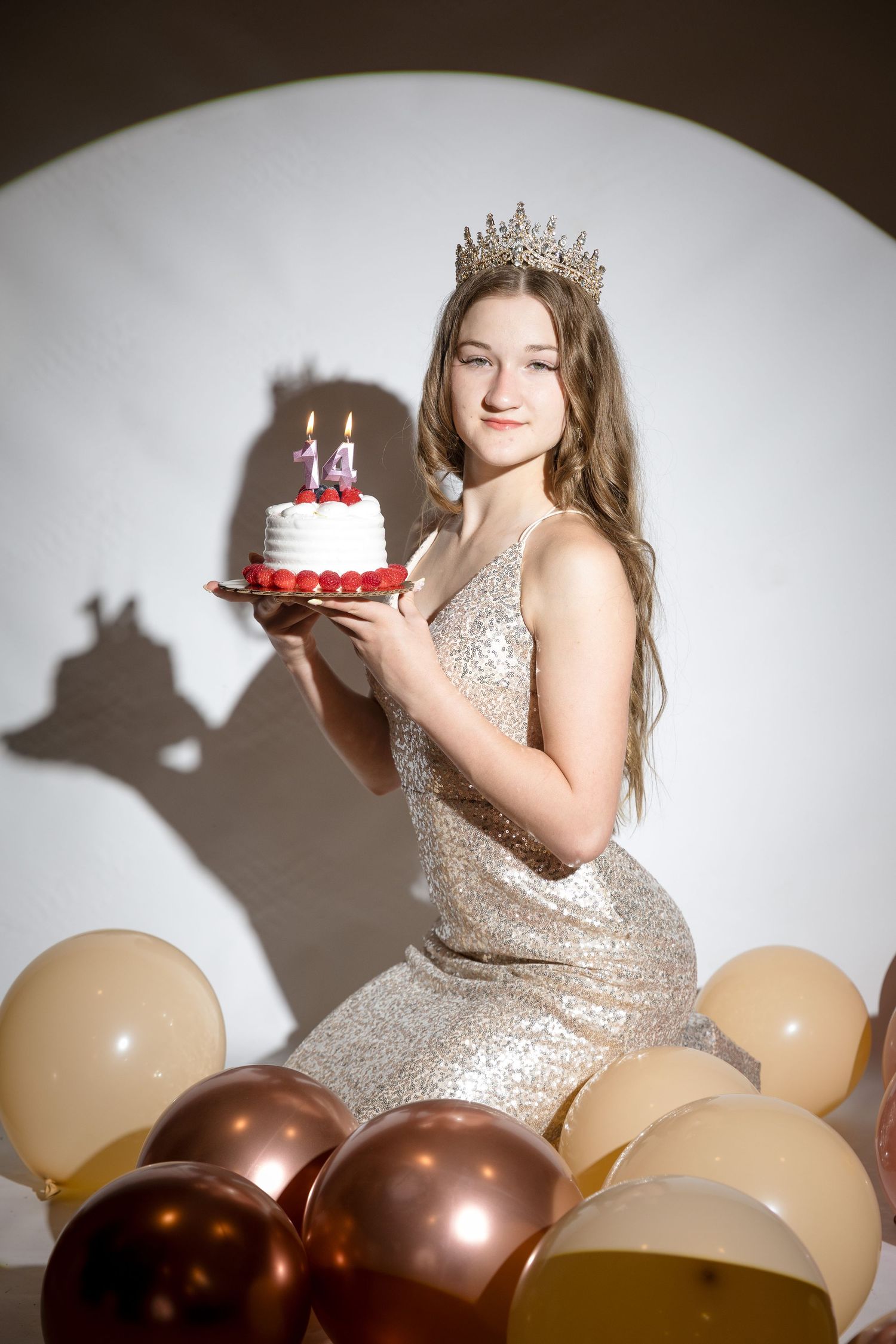 Teen girl in sequin dress and crown holds birthday cake with candles, surrounded by gold and rose gold balloons.