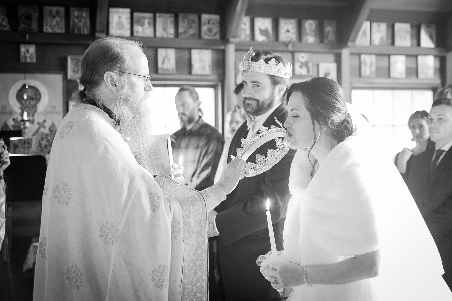 Orthodox priest places crown on bride during traditional wedding ceremony while groom watches holding candle.