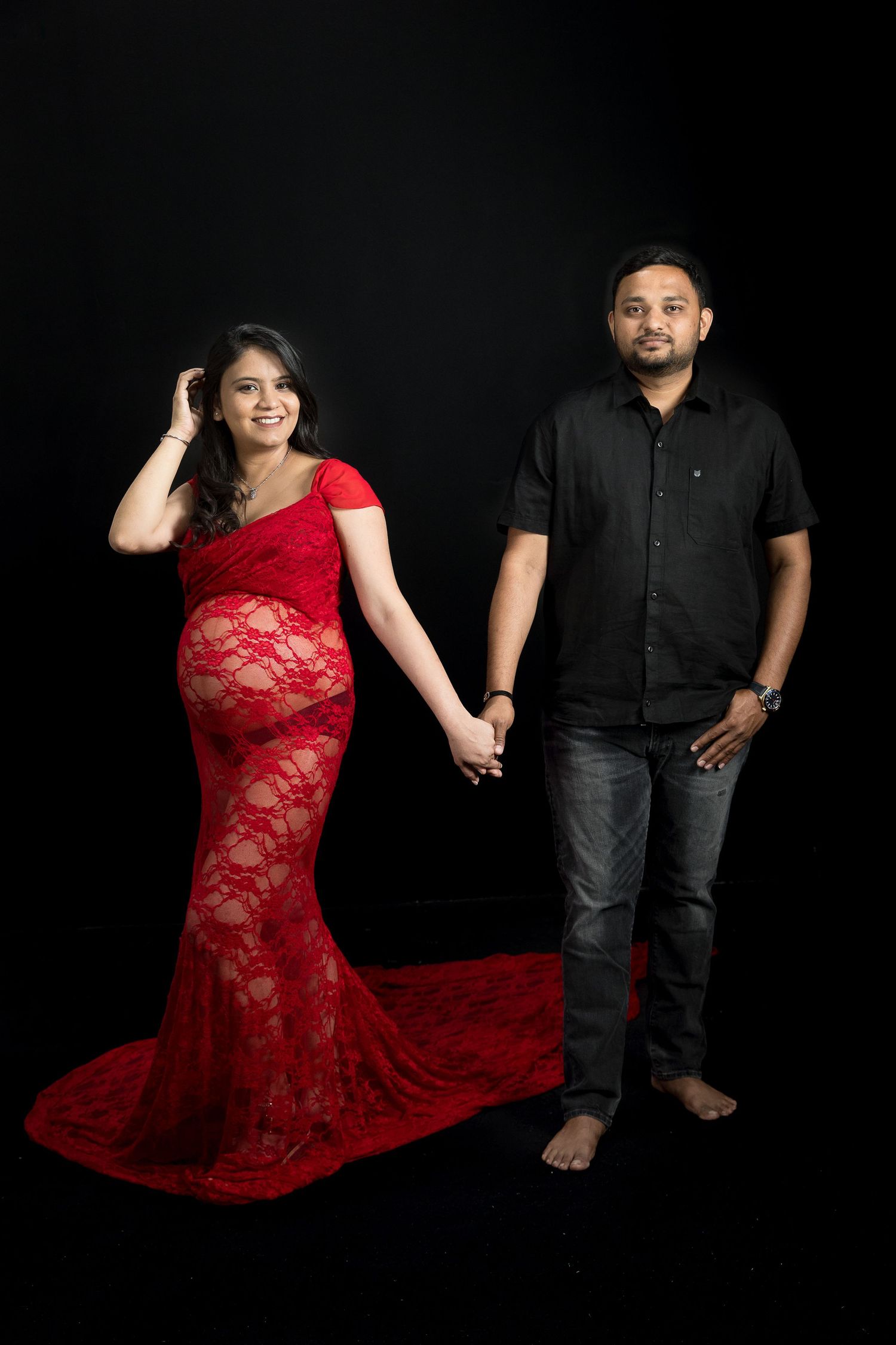 Pregnant woman in red lace gown holds hands with man in black shirt and jeans against dark studio background.
