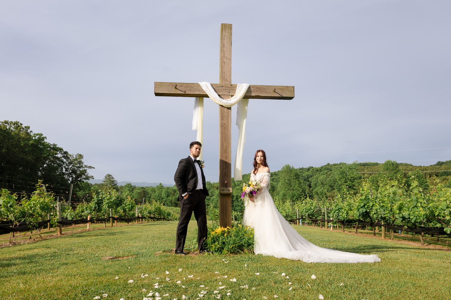 Bride and groom posing before a large wooden cross adorned with white fabric in a lush vineyard setting.