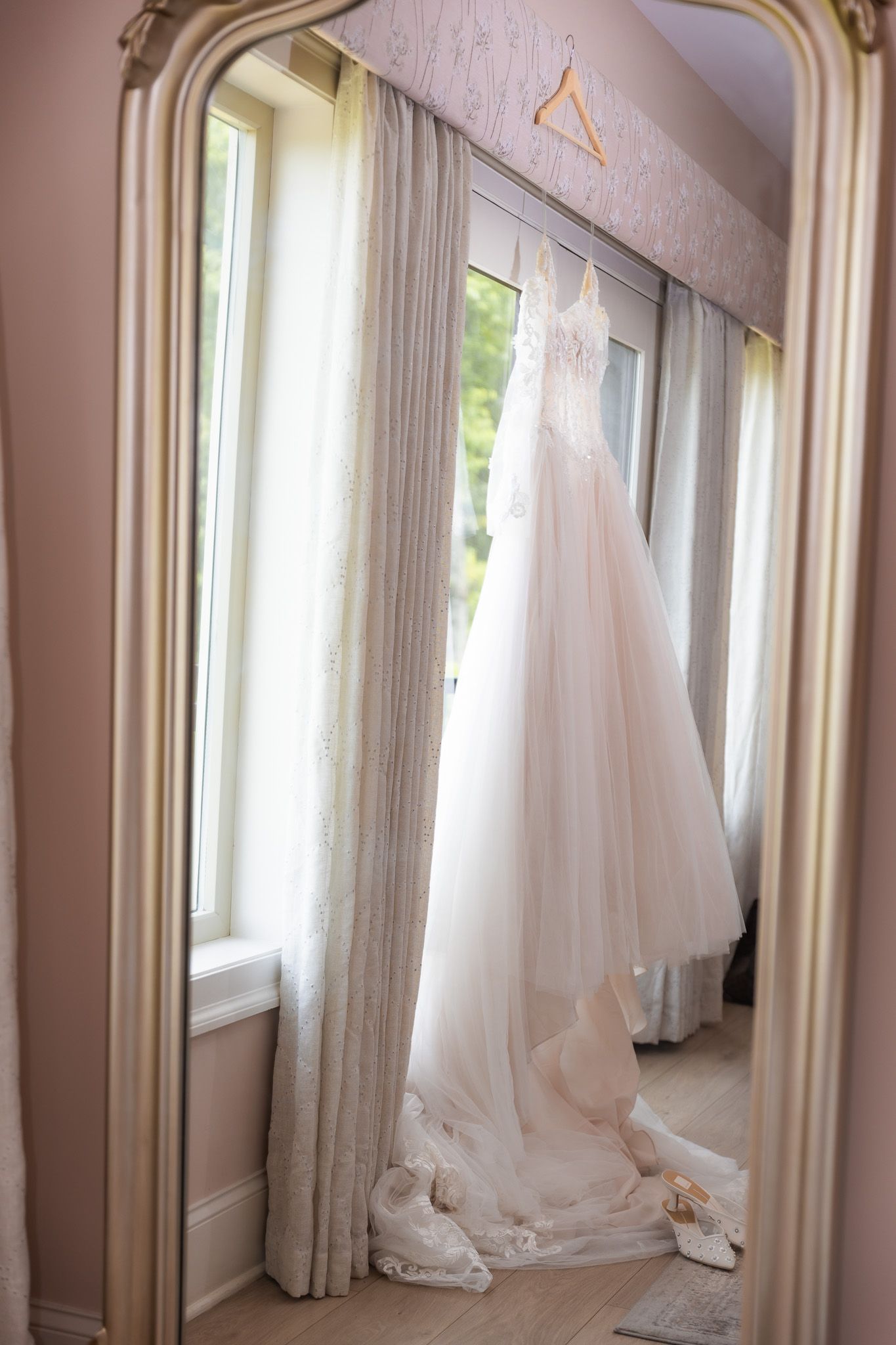 Elegant white wedding gown hanging in a ornate gold mirror, reflecting soft natural light in a bridal suite.
