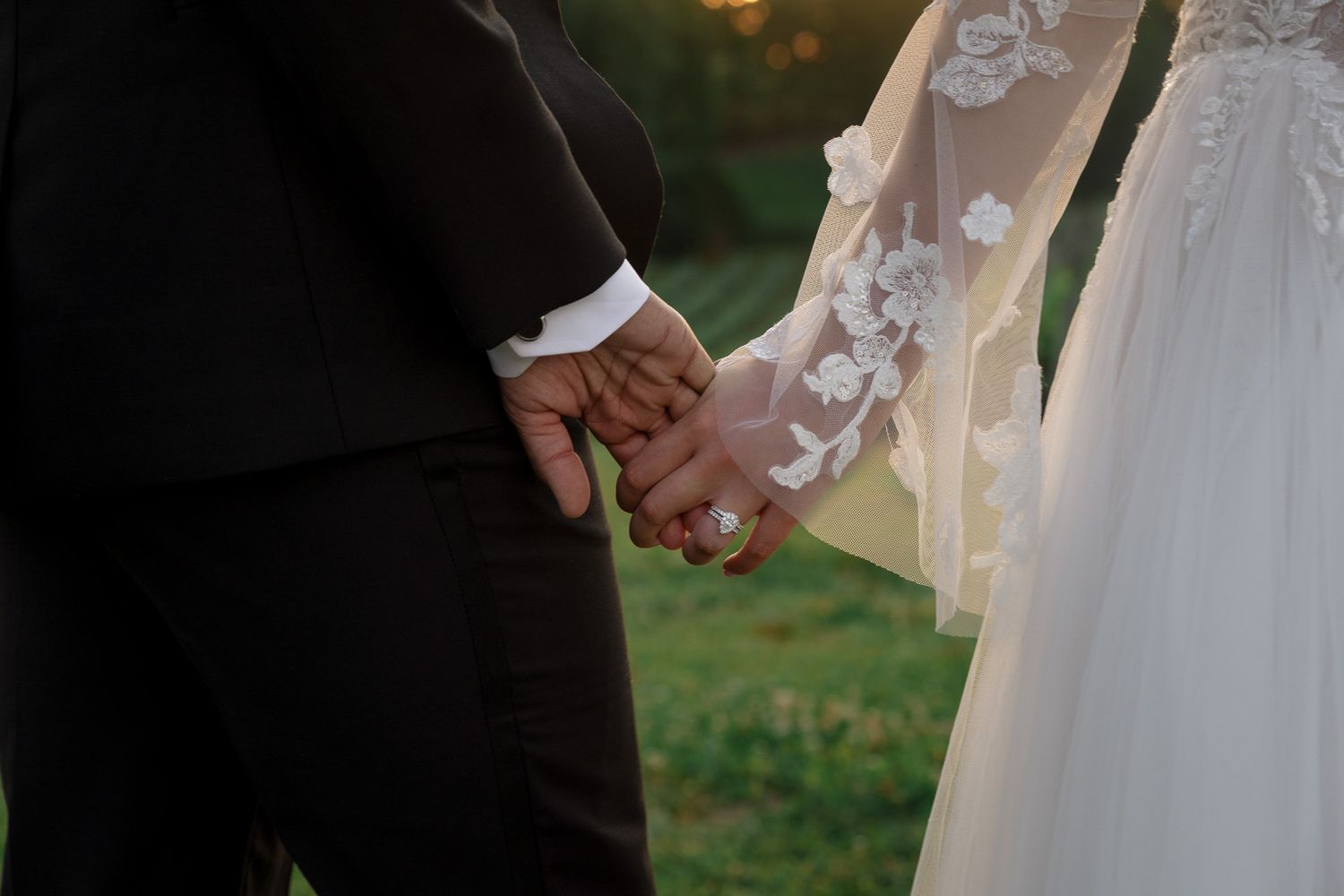 Bride and groom holding hands outdoors, showcasing wedding rings and elegant lace bridal gown with floral details.