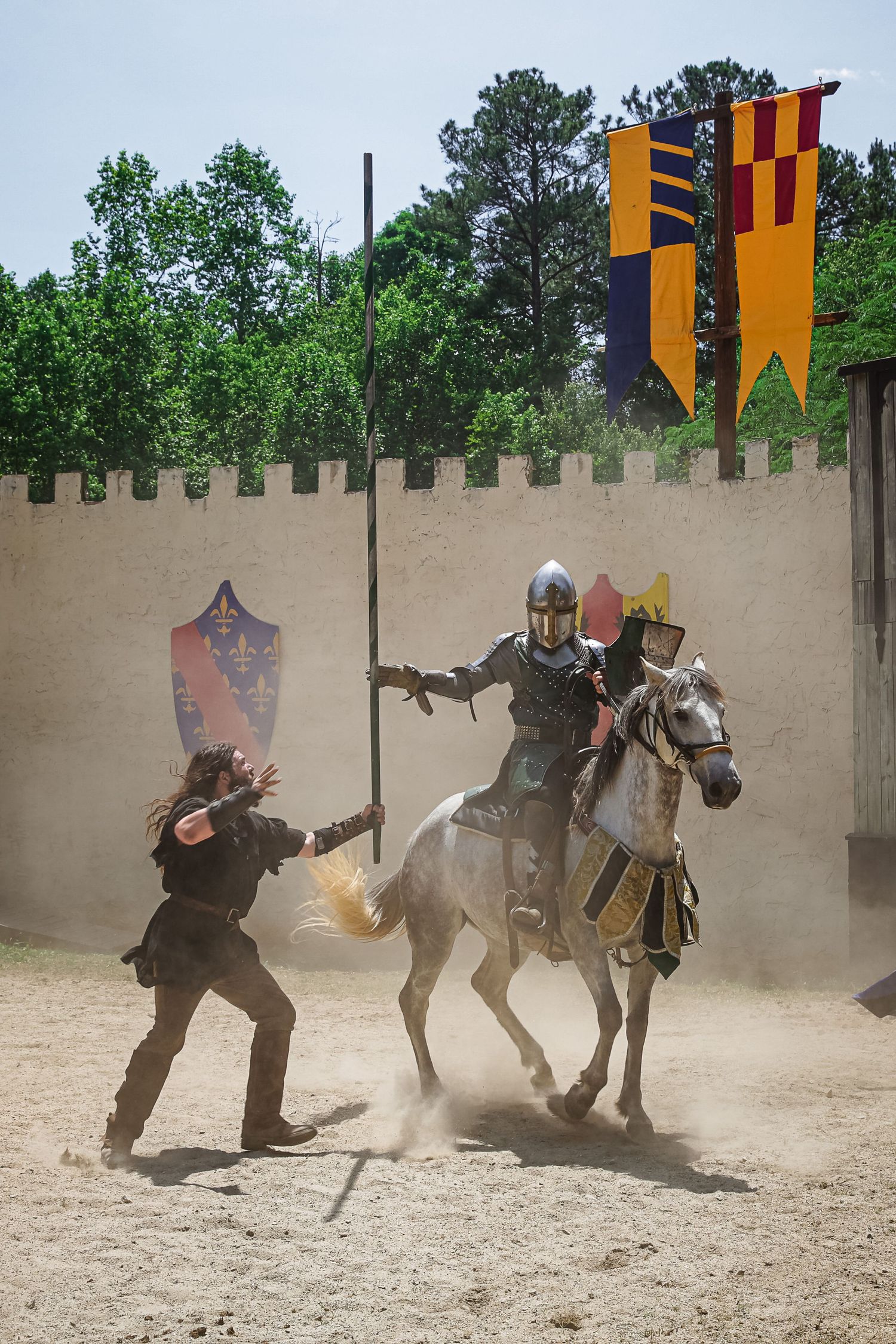 Medieval combat demonstration with armored fighters and horses kicking up dust in a castle arena.