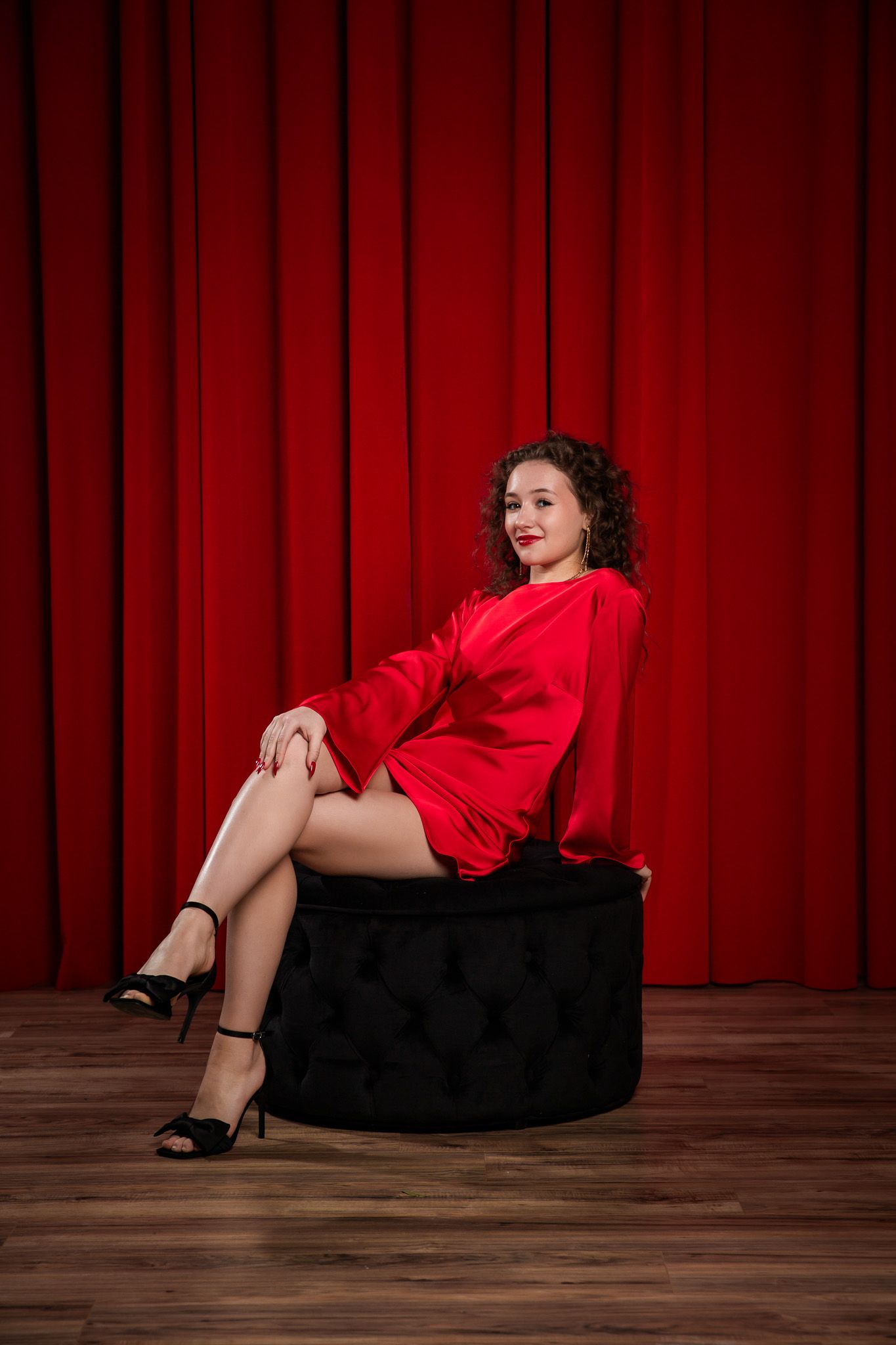 Artistic portrait with red dress against theatrical red curtain and black ottoman.