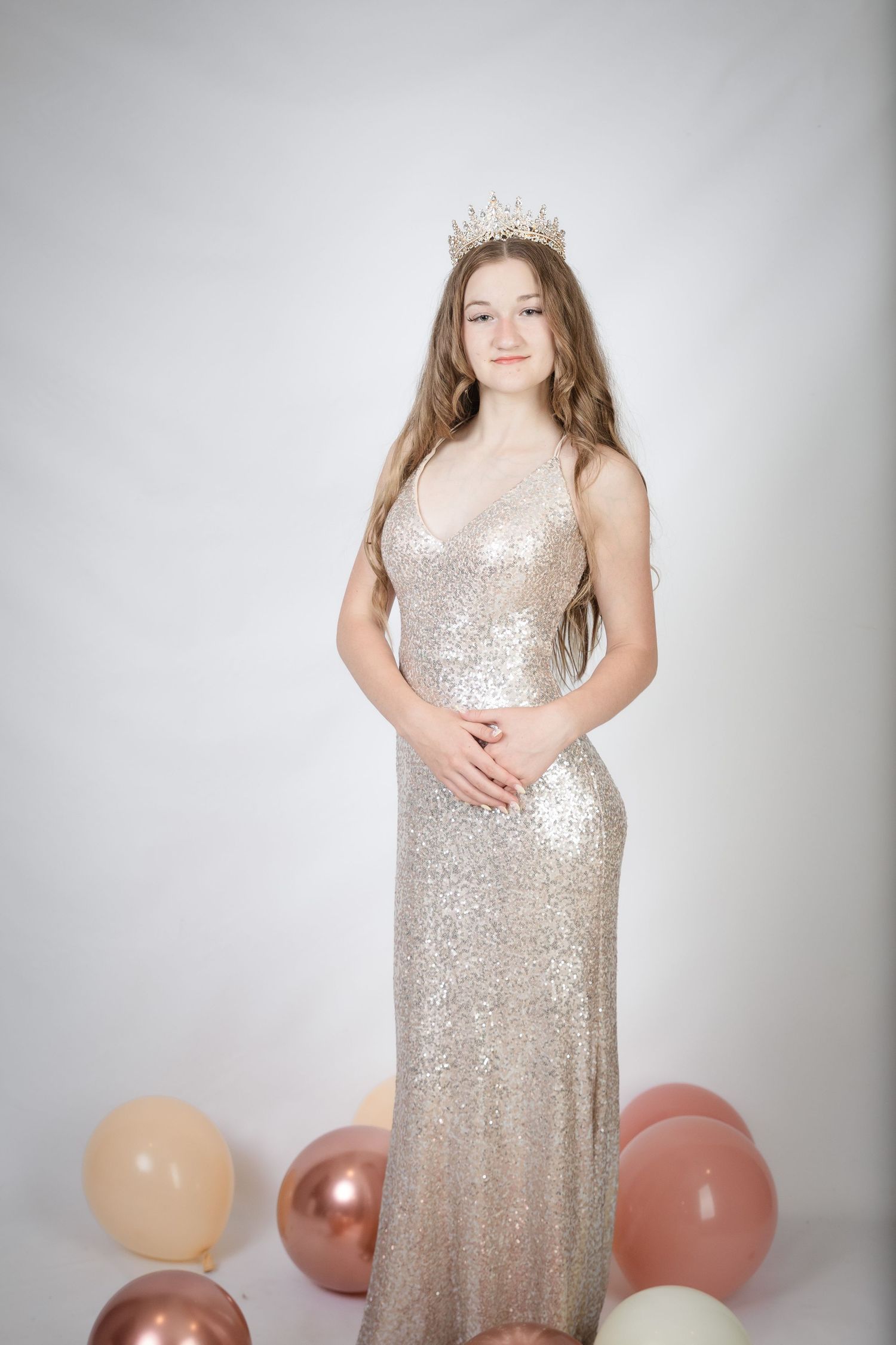 Teen girl in gold sequin gown and crystal crown poses with rose gold balloons against white background.
