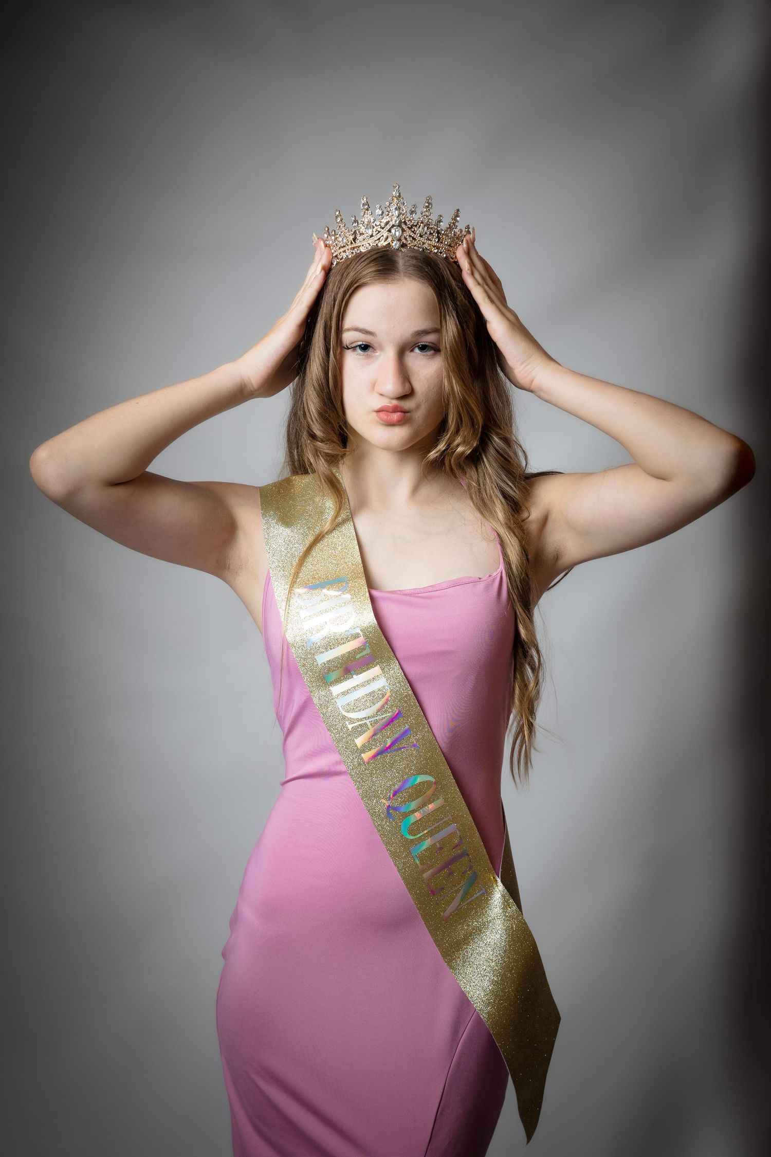 Young woman in pink dress wearing gold crown and birthday sash, posing for a birthday celebration portrait.