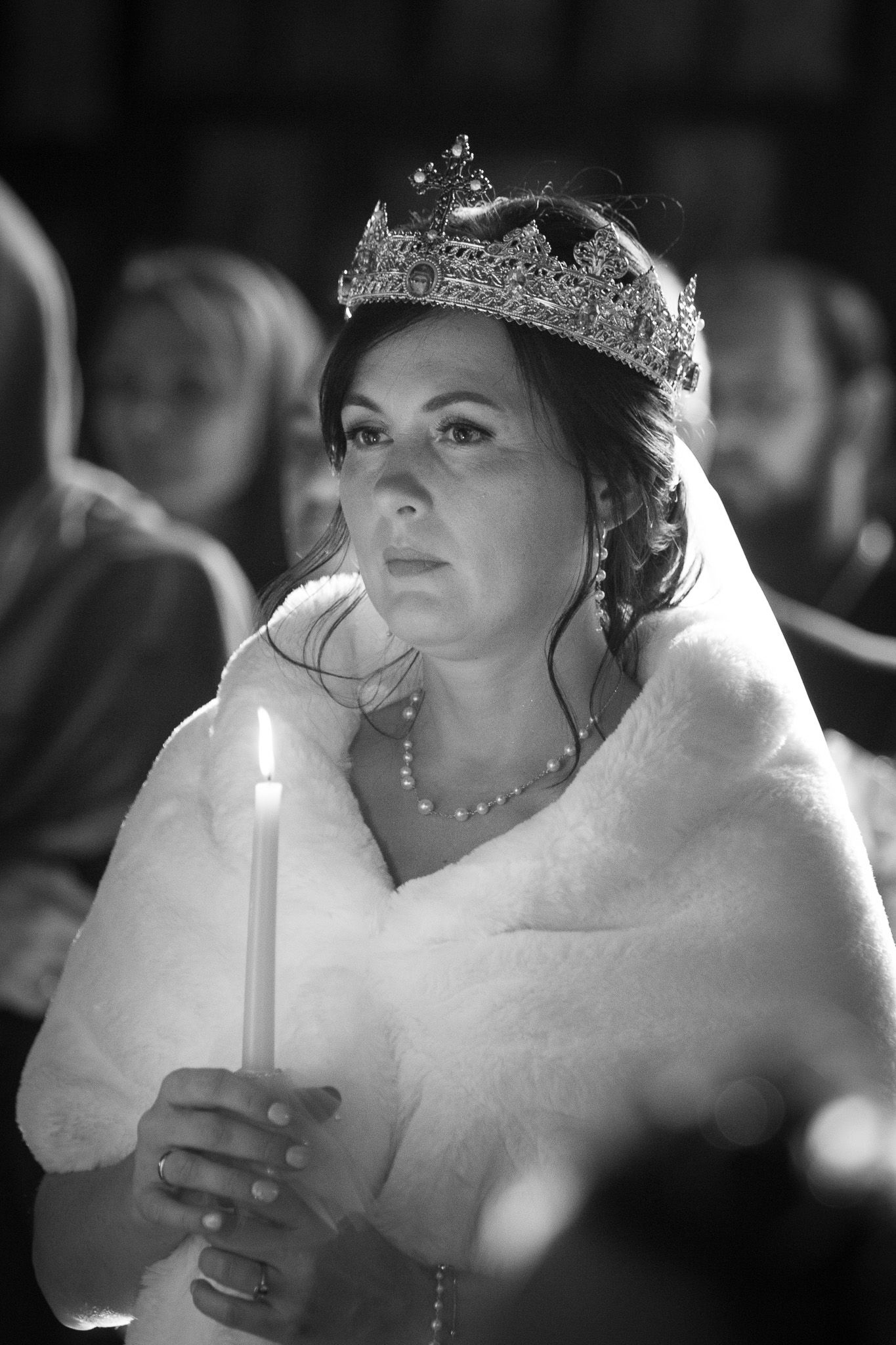 Black and white photo of a bride wearing a tiara and fur stole, holding a candle during a wedding ceremony.