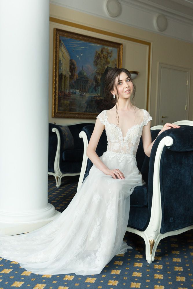 Elegant bride in lace wedding gown poses gracefully beside a velvet chair in a luxurious hotel suite.