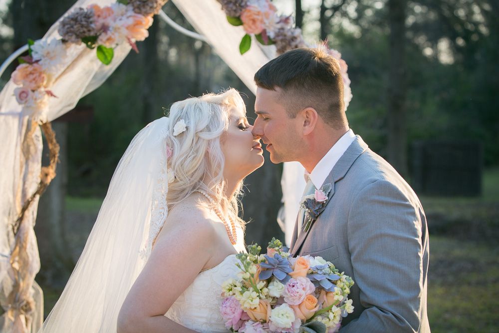 Bride and groom share first kiss under floral arch at outdoor wedding ceremony.
