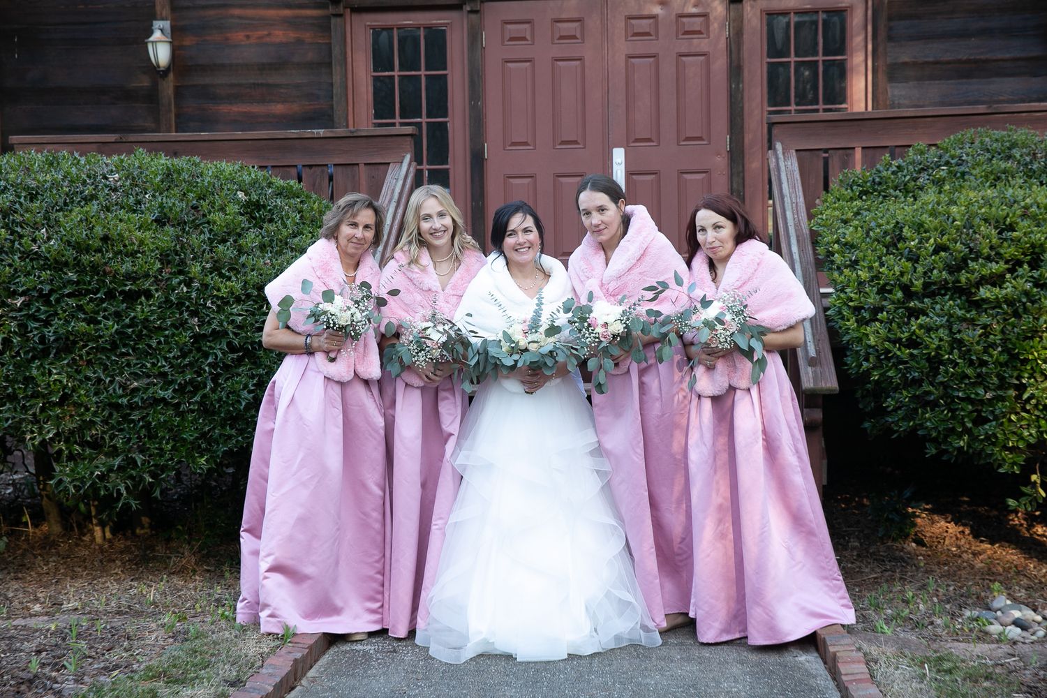 Bride in white gown poses with four bridesmaids in pink dresses holding bouquets outside a rustic wooden building.