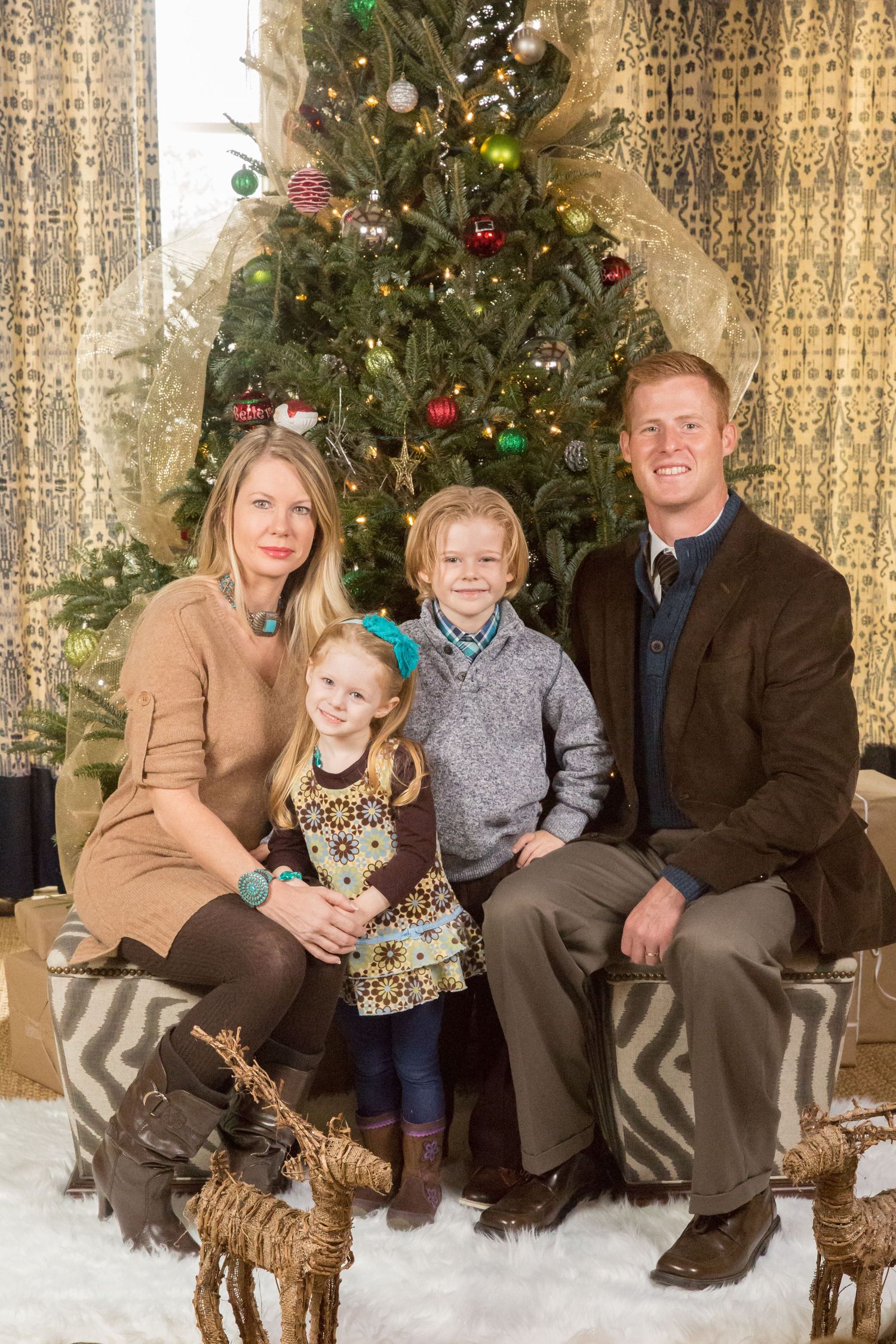 Family seated on rustic bench with Christmas tree and golden curtain backdrop for holiday portraits.