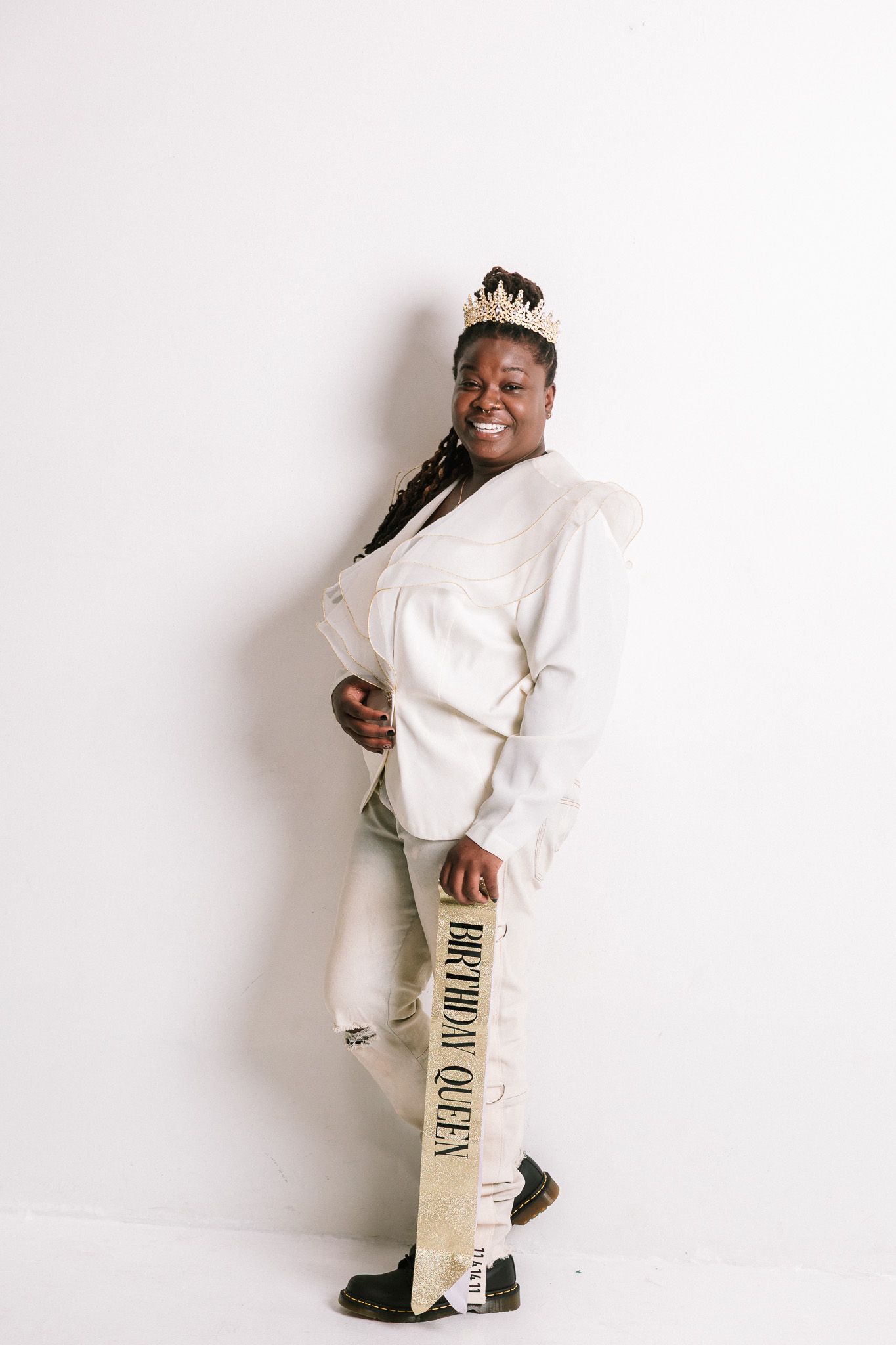 Smiling woman wearing a crown and Birthday Queen sash, dressed in white outfit, posing against a white background.