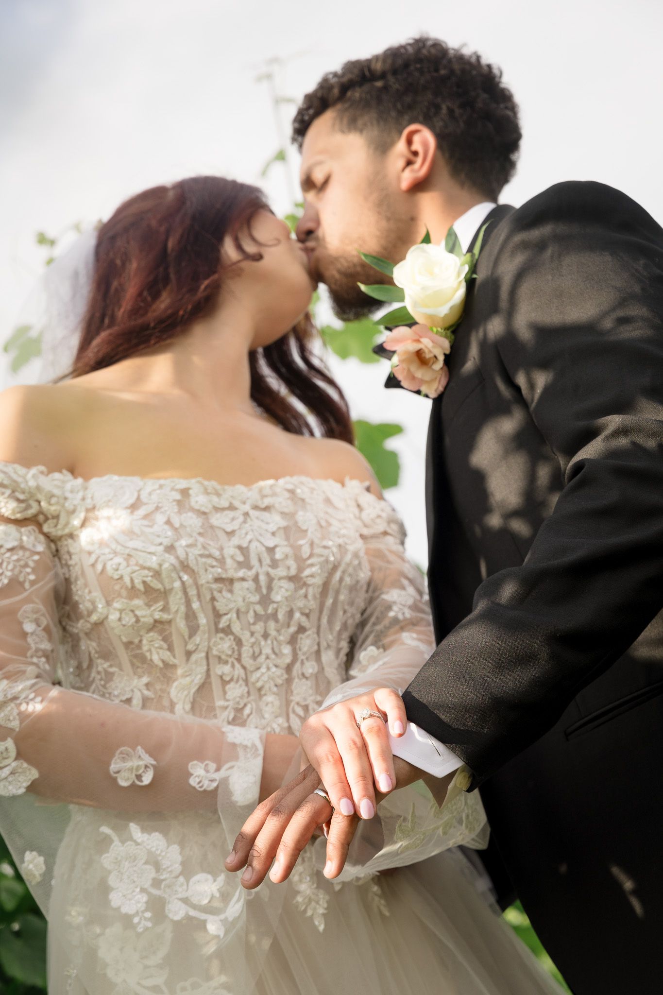 Bride and groom sharing a kiss at their wedding, bride in lace off-shoulder gown, groom in black suit with white boutonniere.