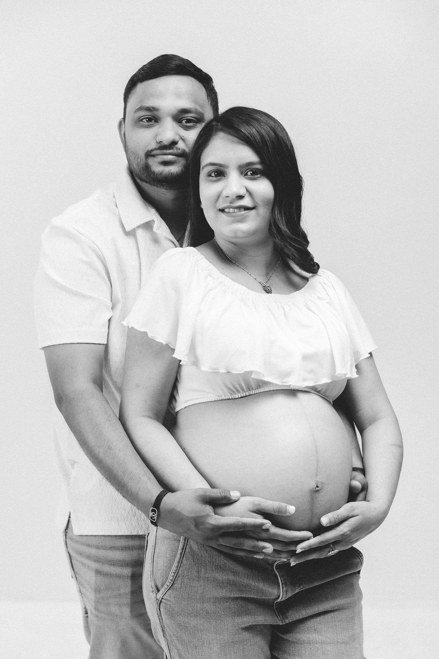 Black and white photo of a pregnant woman and her partner embracing, both smiling, holding her baby bump.