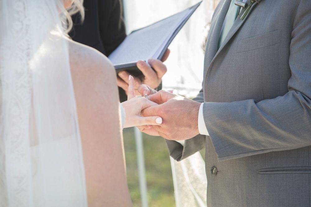 Groom placing wedding ring on bride's finger during outdoor ceremony.