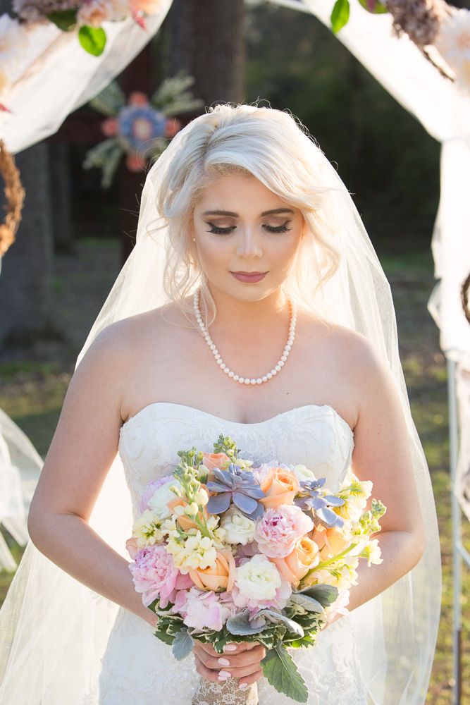 Bride in white strapless gown with veil and pearl necklace holds colorful bouquet of pastel flowers outdoors.
