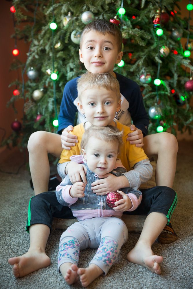 Siblings sitting together on floor in front of lit Christmas tree during holiday photo session.