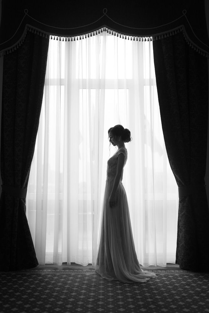 Black and white silhouette of a bride in a wedding gown standing before a large curtained window.