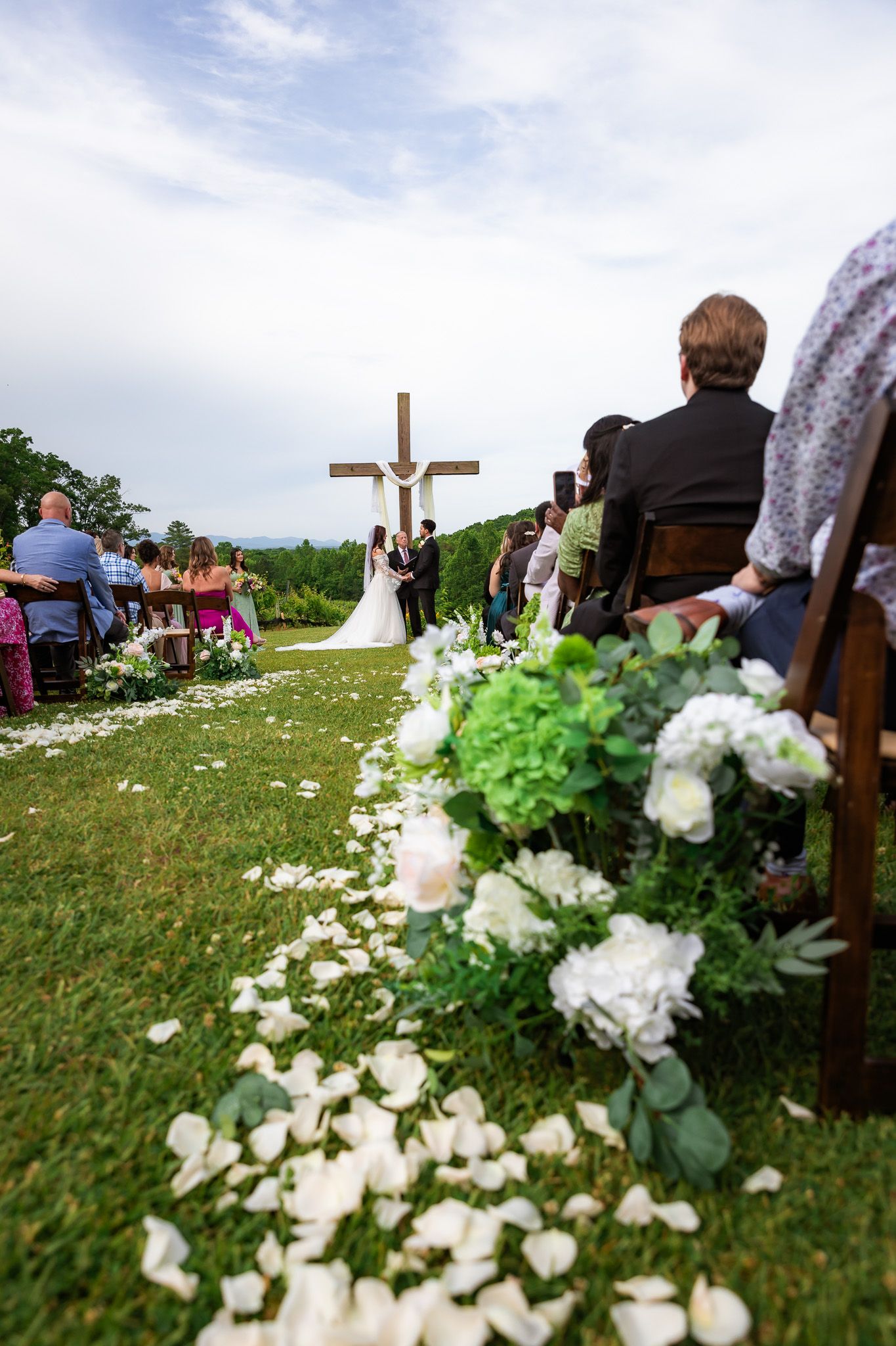 Outdoor wedding ceremony on green grass with white rose petal aisle, wooden cross altar, and guests seated in wooden chairs.