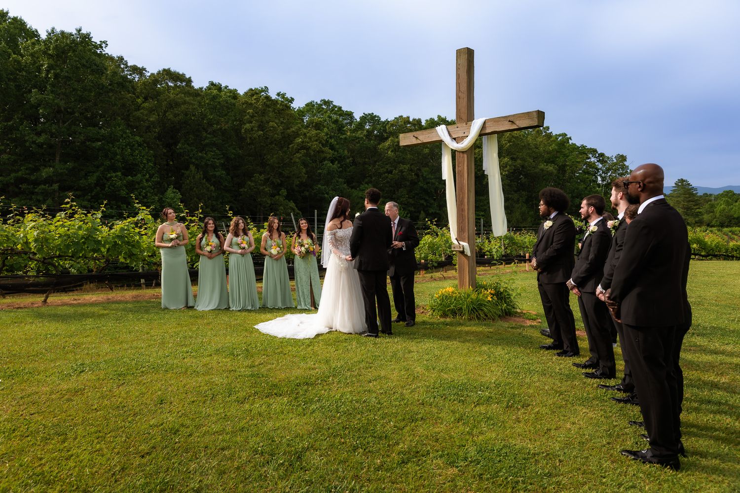 Outdoor wedding ceremony with bridal party before a wooden cross, bridesmaids in sage green gowns, groomsmen in black suits.
