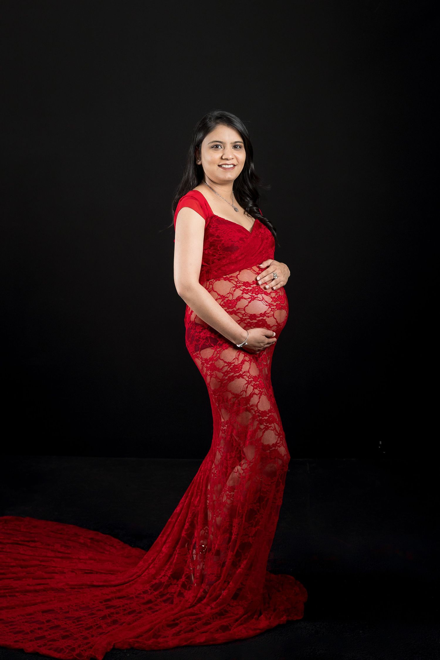 Pregnant woman in elegant red lace gown posing gracefully against black background in professional maternity photo shoot.