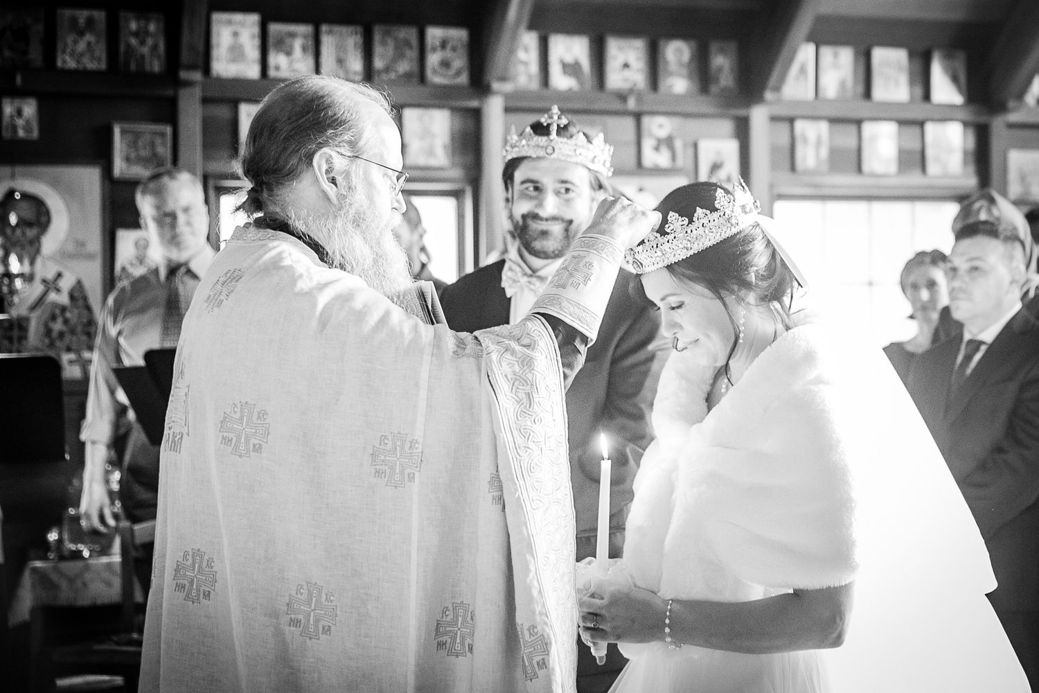 Black and white Orthodox wedding ceremony with priest placing crown on bride while groom smiles in a decorated church.