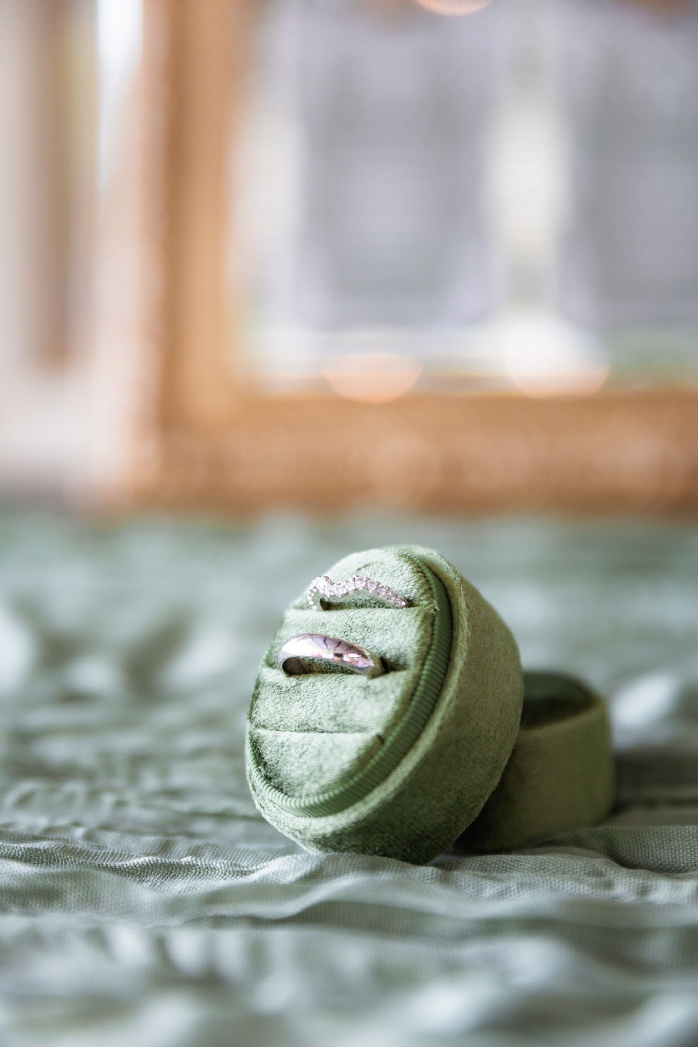 Two silver wedding bands displayed in an open round green velvet ring box on a sage green fabric surface.