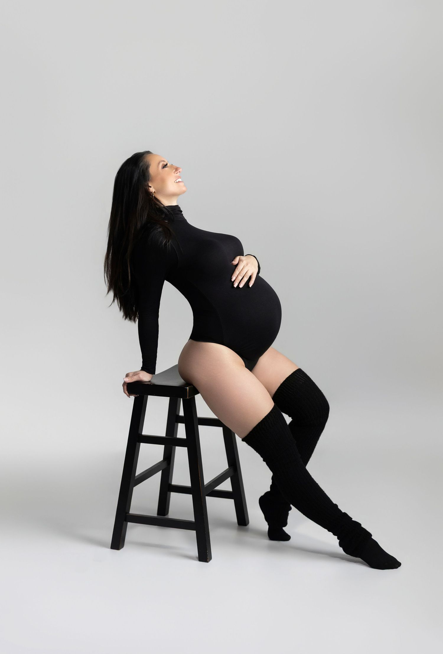 Artistic maternity poses on black stool showcasing pregnant figure in dark outfit.