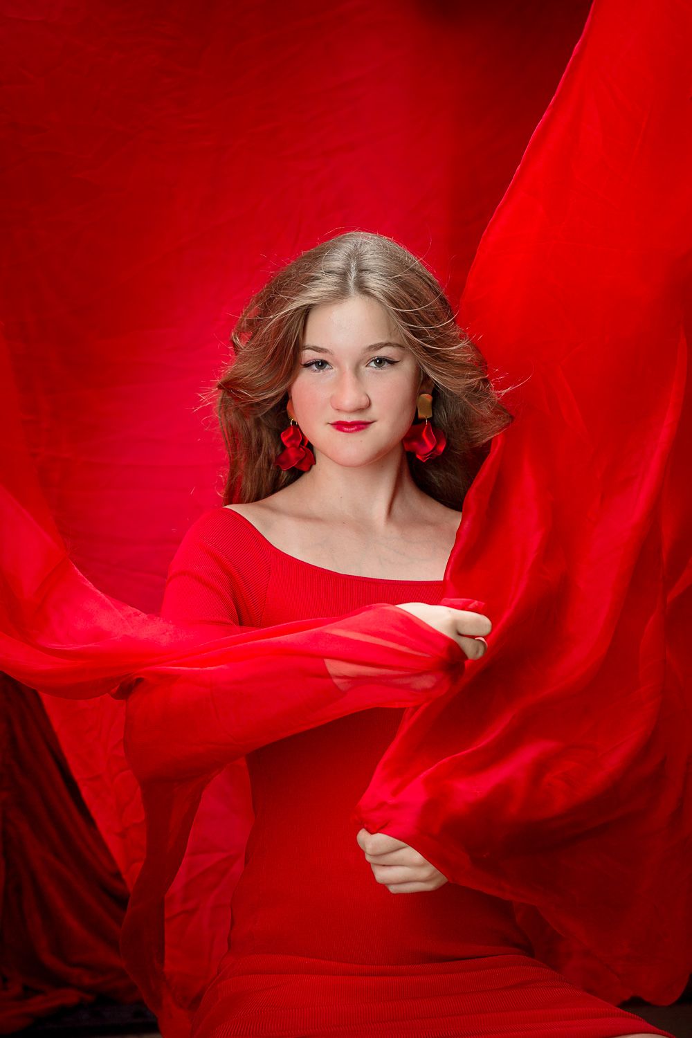 Young woman in red dress surrounded by flowing red fabric against red background, wearing gold earrings and red lipstick.
