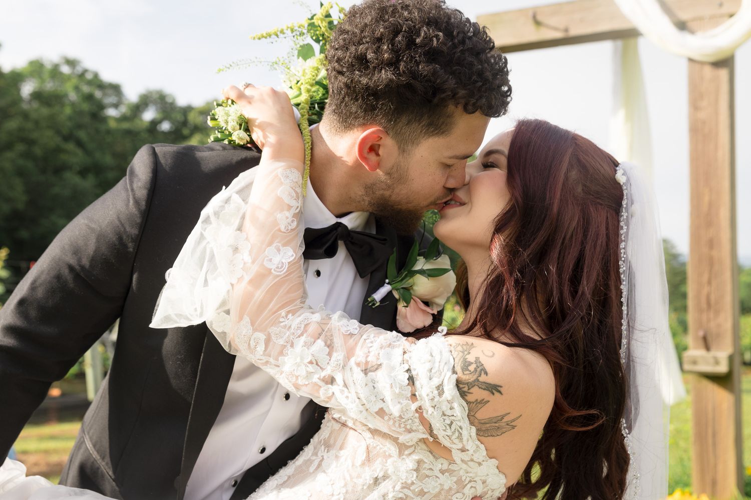 A bride and groom share a romantic kiss at their outdoor wedding ceremony, with the bride wearing lace and the groom in a tuxedo.
