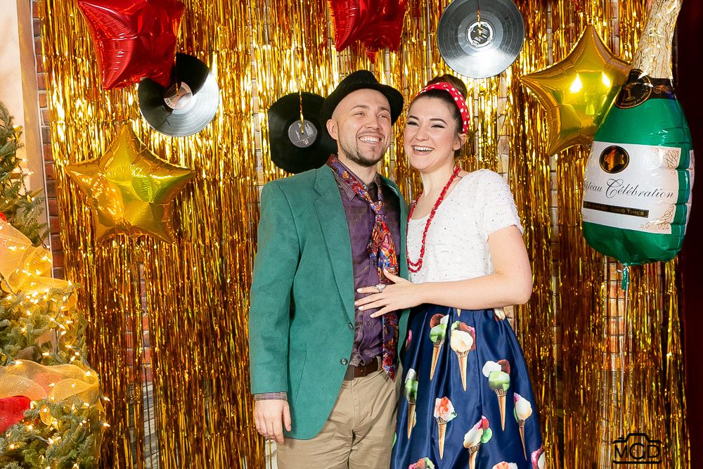 Couple posing for holiday photos in front of gold tinsel backdrop with red star balloons.