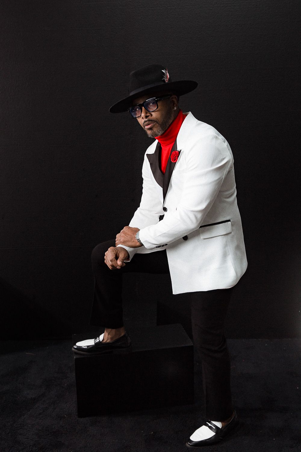 Person in white suit jacket and fedora strikes dramatic pose on black backdrop.