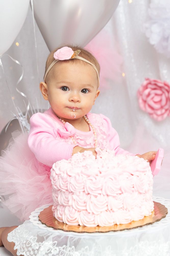 Baby girl in pink tutu celebrates first birthday with a pink rosette smash cake, balloons in background.