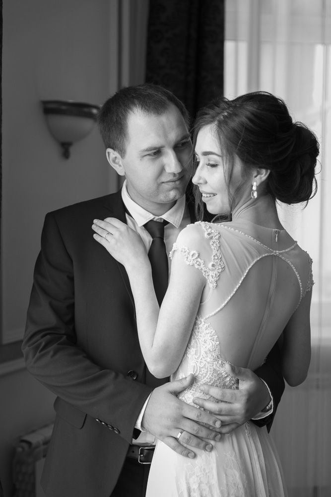 Black and white photo of a groom embracing his bride in a tender wedding moment indoors.