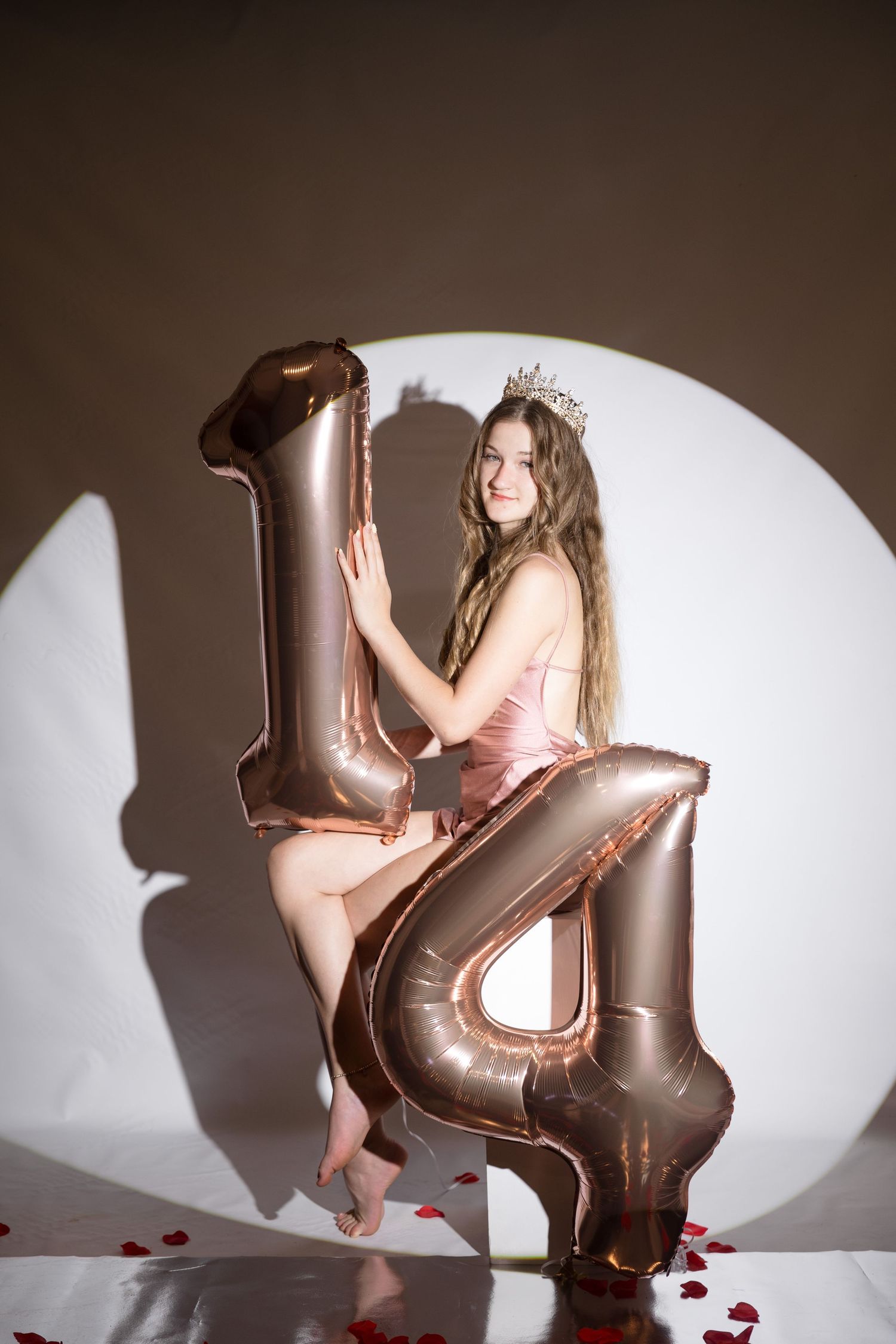Young girl in pink dress and crown poses with large number 14 balloons for birthday celebration.