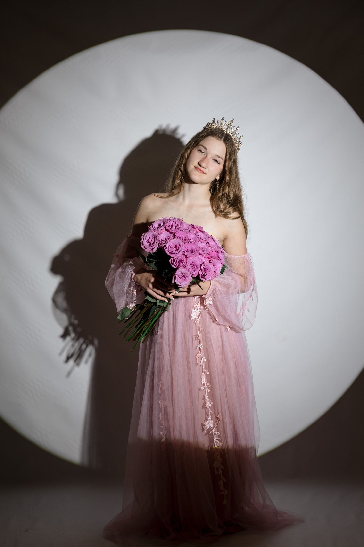 Young woman in pink tulle gown and crown holds bouquet of pink roses, posing elegantly in studio spotlight.