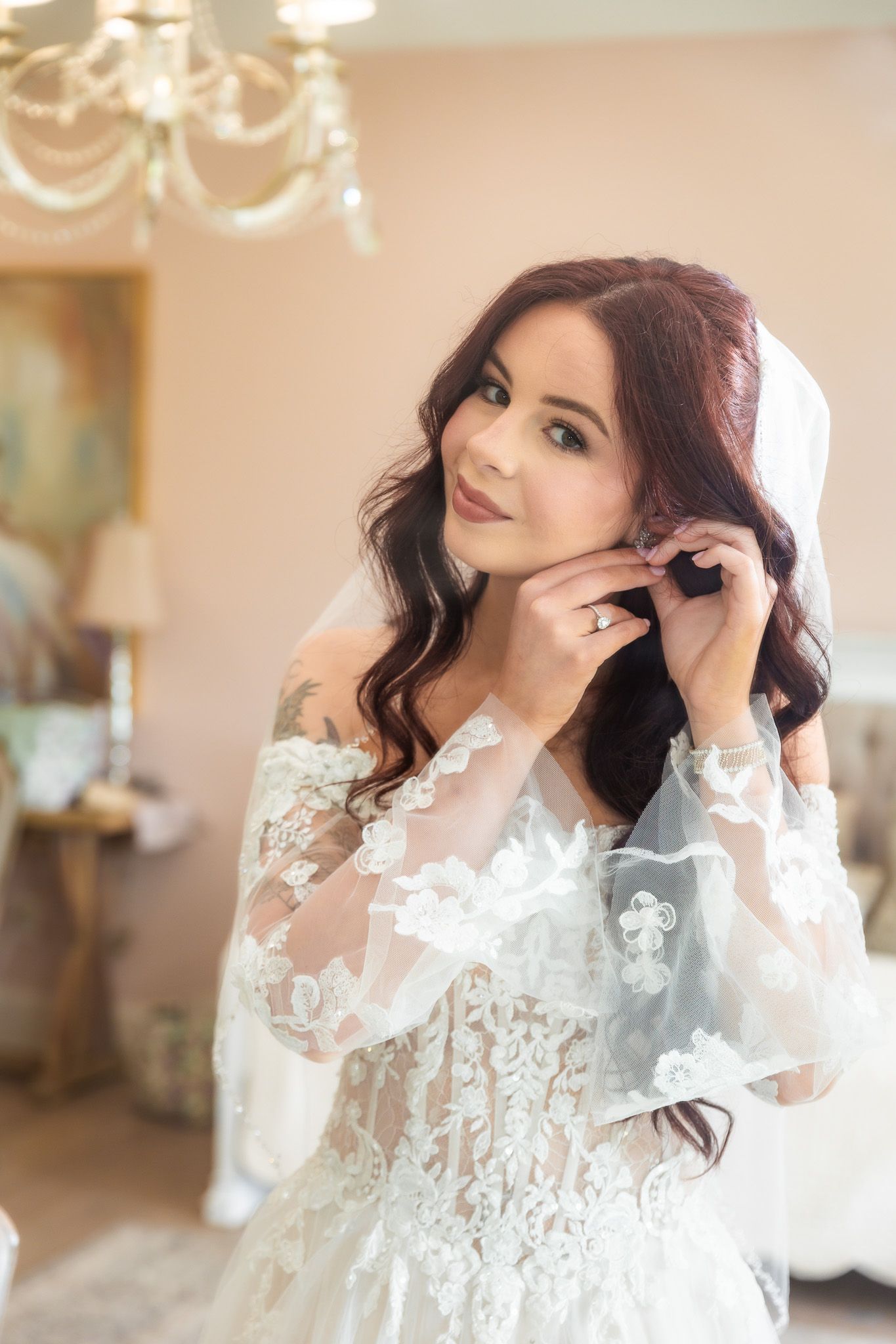 Bride in lace wedding gown adjusting earring while wearing veil indoors.